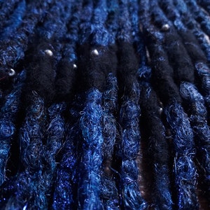 Könnte beinhalten: Nahaufnahme von blauen und schwarzen synthetischen Dreadlocks mit silbernen Perlen. Die Dreadlocks sind in Reihen angeordnet und haben eine leicht strukturierte Oberfläche.