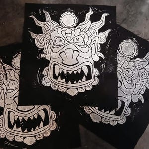 May include: Three black and white linocut prints of a stylized mask with sharp teeth and ornate details. The prints are arranged on a dark surface, showcasing the bold contrast of the design.