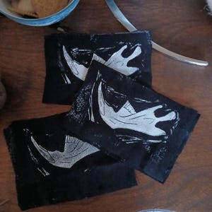 Pode incluir: Quatro bases quadradas pretas com um design de osso branco estilizado. As bases estão dispostas sobre uma superfície de madeira. Uma tigela azul e um utensílio de servir prateado estão no fundo.