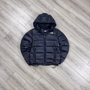 May include: A dark navy blue puffer jacket with a hood, featuring horizontal quilted stitching, a full-length front zipper, and the white The North Face logo embroidered on the upper left chest and back right shoulder.