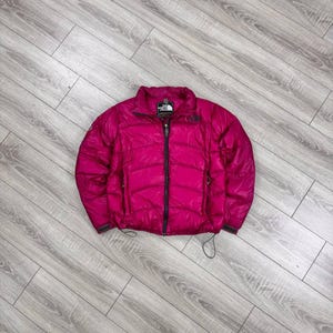 May include: A bright magenta puffer jacket featuring a high collar, a full-length black front zipper, and horizontal quilted stitching. The North Face logo is embroidered on the upper left chest, and the jacket includes adjustable drawstrings.