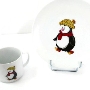 Könnte beinhalten: Ein weißes Keramik-Geschirrset für Kinder mit einem Pinguin-Motiv. Der Teller zeigt einen Pinguin mit einem gelben Hut und einem roten Schal. Die Schüssel hat einen Pinguin mit einem gelben Hut.