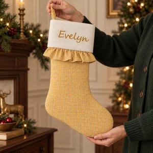 May include: A yellow and white gingham Christmas stocking featuring a white cuff with the name Evelyn embroidered in gold thread. The stocking includes a ruffled fabric trim below the cuff and a small loop at the top for hanging.