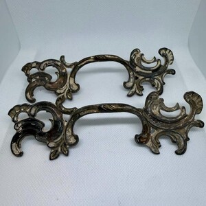 May include: Two matching metal drawer pulls featuring an ornate, scrolling design with a distressed finish. The hardware displays a dark, tarnished metallic base with contrasting white, weathered paint accents along the intricate, curved edges.