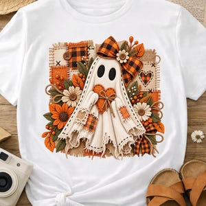 May include: A white t-shirt featuring a graphic of a ghost made of fabric patches, holding an orange heart. The design includes a plaid bow, sunflowers, daisies, and autumn leaves, set against a background of textured burlap and plaid squares.