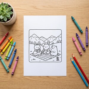 K&ouml;nnte beinhalten: Ein wei&szlig;es Blatt Papier mit einer schwarz-wei&szlig;en Strichzeichnung von zwei Kaninchen und einem Hamster, die ein Picknick auf einer karierten Decke vor Bergen machen, umgeben von bunten Wachsmalstiften und einer kleinen gr&uuml;nen Sukkulente.
