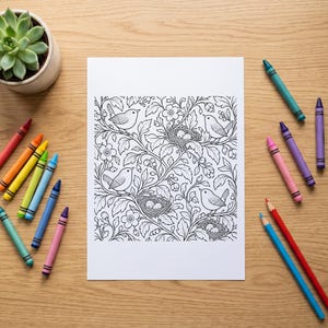 May include: A white rectangular sheet of paper featuring a detailed black and white line art illustration of birds, nests with eggs, flowers, and leafy branches, surrounded by an assortment of colorful wax crayons on a light wood surface.