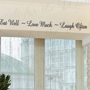 May include: Black vinyl wall decal featuring the phrase "Eat Well ~ Love Much ~ Laugh Often" in a cursive font. The decal is positioned above a window with sheer curtains. The words are a decorative element for home decor, suitable for any room.