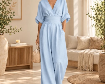 May include: A light blue jumpsuit featuring a V-neckline, elbow-length sleeves with folded cuffs, and a wide-leg silhouette. The garment includes a fitted waistband and is paired with a small, textured, light brown woven handbag.