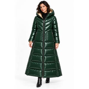 Può includere: Un lungo piumino verde scuro lucido con chiusura a zip frontale, cappuccio con fodera interna chiara e impunture orizzontali. Il cappotto arriva fino alle caviglie e presenta due tasche laterali sul davanti.