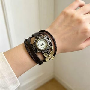 May include: A dark brown leather wrap watch featuring a round analog face with black numerals. The watch case is framed by a decorative metal butterfly design and includes a dangling brass-toned butterfly charm attached to the braided strap.