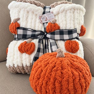 May include: A white chunky knit blanket with small orange pumpkin appliques, tied with a black and white buffalo check ribbon. A large, round, orange crocheted pumpkin with a tan stem sits in front of the folded blanket.
