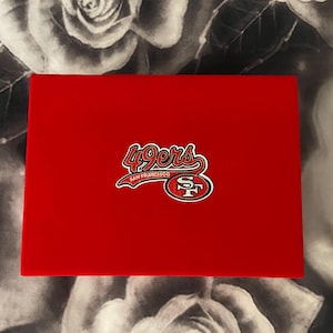May include: A rectangular red box featuring the San Francisco 49ers logo centered on the lid. The logo displays the text 49ers in a stylized red font with a white outline, positioned above the letters SF enclosed in a red oval with a white border.