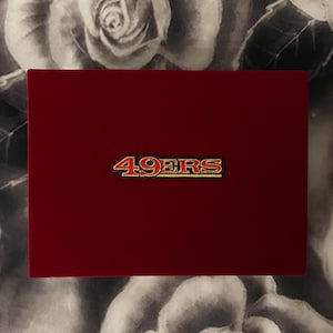 May include: A rectangular, deep burgundy velvet box featuring the word 49ERS embroidered in red and gold thread centered on the lid. The surface of the box has a soft, matte texture, and the lettering is outlined in a bold, metallic gold tone.