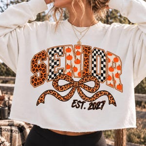 Peut inclure: Un sweat-shirt blanc à manches longues avec le mot SENIOR en grandes lettres majuscules à motifs léopard, damier noir et blanc et citrouilles orange. Un nœud léopard est placé sous le texte avec l'inscription EST. 2027.