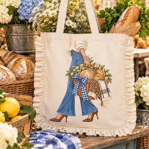 May include: A cream-colored canvas tote bag with ruffled side edges, featuring a graphic print of legs in blue flared jeans and leopard print heels, carrying a woven basket filled with bread, daisies, and a blue gingham bow.