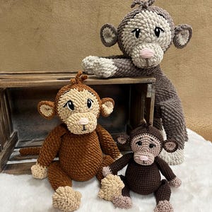May include: Three handmade crochet monkey dolls in shades of brown and beige. The monkeys have detailed features, including expressive faces, ears, and limbs. One monkey is perched on a wooden crate, while the others sit on a white surface.