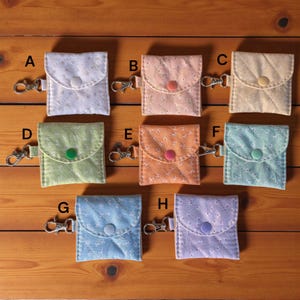 May include: Eight small, quilted fabric pouches in various pastel colors, each with a floral pattern and a snap closure. Each pouch has a silver-colored metal clasp attached. The pouches are arranged on a wooden surface.