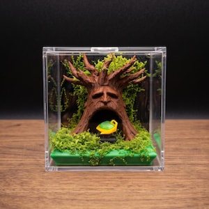 May include: A clear square display case containing a brown sculpted tree with a face and a hollow trunk. Inside the trunk sits a small yellow and green object. The base is covered in bright green moss, and the tree is surrounded by green foliage.