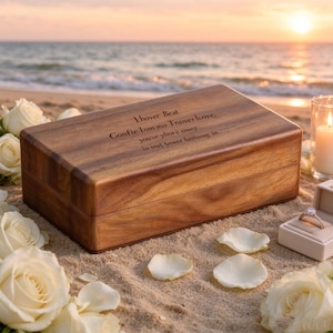 May include: A rectangular wooden box with a dark brown grain, featuring an engraved inscription on the lid that reads "I hover Beat Gonfie Iom mo Traiver Icove, youve ybor'c owes... iw inat yower entiong in...". White roses and a ring box are nearby.