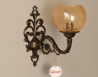 Puede incluir: Un aplique de pared con una placa metálica decorativa en tono bronce oscuro con detalles de volutas y un brazo curvo. Sostiene una pantalla esférica de vidrio craquelado color ámbar. Una etiqueta redonda blanca muestra el texto Oneels Midcentury.