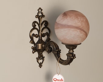 Puede incluir: Un aplique de pared con una estructura de metal en tono bronce oscuro con volutas intrincadas y una pantalla esférica de vidrio veteado en tonos rosa suave y beige. Una pequeña etiqueta redonda con el texto Oneels Midcentury cuelga debajo.