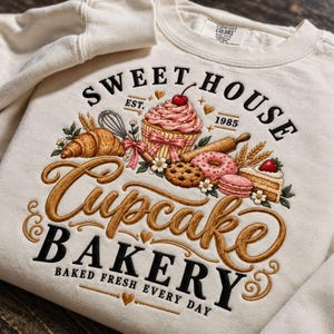 Puede incluir: Una sudadera de cuello redondo color crema con un estampado gráfico que incluye el texto "SWEET HOUSE EST. 1985" y "Cupcake BAKERY BAKED FRESH EVERY DAY". El diseño muestra ilustraciones de un cupcake, cruasán, dónut, galleta y pastel.