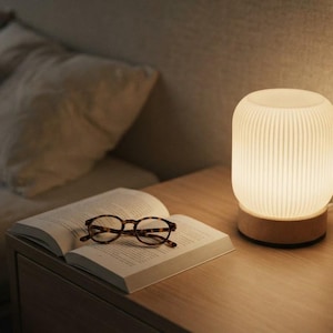 Peut inclure: Une lampe de table cylindrique avec un abat-jour en verre blanc à côtes verticales et une base en bois clair. La lampe émet une lueur chaude et douce. À côté de la lampe se trouve un livre ouvert avec une paire de lunettes à motif écaille.