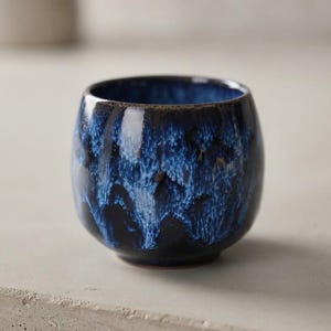 Könnte beinhalten: Eine kleine, abgerundete Keramiktasse mit einer tief dunkelblauen und schwarzen Glasur, die lebhafte, streifige kobaltblaue Akzente aufweist. Die Oberfläche hat ein glänzendes Finish mit einer gesprenkelten Textur und einem glatten, runden Rand.
