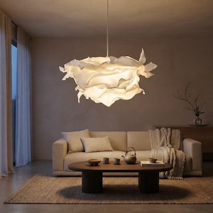 Peut inclure: Une grande suspension lumineuse blanche et sculpturale, composée de panneaux superposés aux formes fluides rappelant du tissu, diffusant une lumière douce. Le luminaire est suspendu au plafond par un cordon fin et éclaire la pièce.