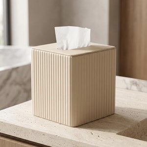 Puede incluir: Una caja para pañuelos cuadrada de color beige claro con una textura acanalada vertical en los cuatro lados. La parte superior presenta una superficie plana con una abertura rectangular central de la que sale un pañuelo blanco.