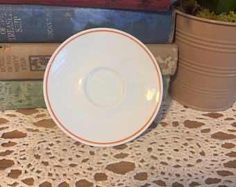 May include: A round white ceramic saucer featuring a thin, vibrant orange circular band near the outer edge. The saucer has a smooth, glossy finish and a central indentation designed to hold a cup securely in place.