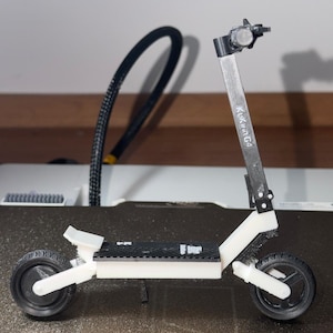 以下が含まれることがあります： 白いフレーム、黒いホイール、斜めのテクスチャラインが入った黒いデッキを備えた電動スクーターのミニチュアモデル。垂直のステアリングコラムは黒と白で、白い文字でKukirin G4と明確にプリントされています。