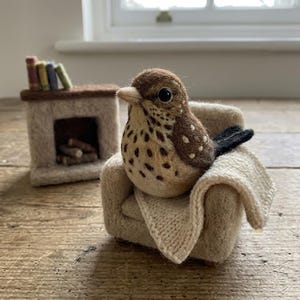 May include: A needle-felted brown and tan bird with dark spots sits in a miniature beige armchair draped with a small knitted blanket. Beside the chair is a tiny felt fireplace containing small logs and topped with four miniature books.