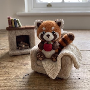 May include: A needle-felted red panda figurine holding a small red apple, sitting in a miniature beige armchair with a cream-coloured knitted blanket. In the background, a small felt fireplace holds tiny books and logs.
