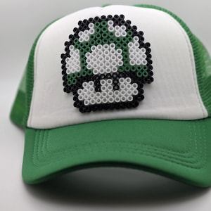 May include: A green and white trucker hat featuring a pixelated mushroom design on the front panel. The mushroom is constructed from small, circular beads in shades of green, white, and black, creating a distinct, textured graphic element.