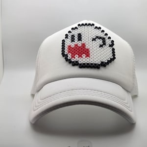 May include: A white baseball cap featuring a pixelated ghost character design on the front panel. The character is constructed from small, circular beads in white, black, and red, creating a distinct, blocky, and textured graphic appearance.