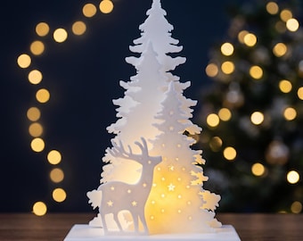 May include: A white, illuminated decorative tabletop ornament featuring a layered pine tree silhouette with a standing deer in the foreground. The structure includes cut-out star shapes that emit a warm, soft glow from within.