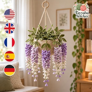 Op de afbeelding: Een gehaakte hangplant met een beige mand gevuld met groene bladeren en trapsgewijze blauweregenbloemen in tinten paars, lavendel en wit. De plant hangt aan een crèmekleurig koord dat is bevestigd aan een houten ring aan de bovenkant.