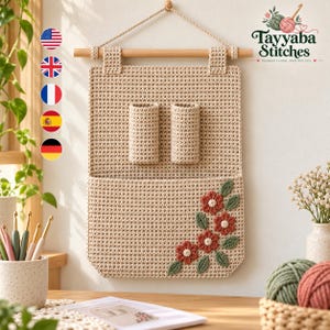May include: A beige crochet wall hanging organizer featuring two vertical cylindrical pockets and a large bottom pocket decorated with four red flowers and green leaves. The organizer is suspended from a wooden dowel with a rope.