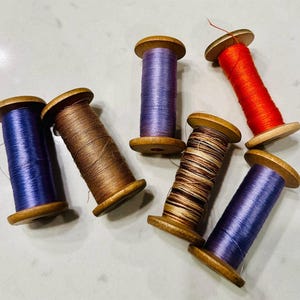 May include: Six wooden spools of thread in various colors including blue, brown, purple, orange, and a variegated brown and black blend. Each spool features a central wooden core with circular ends, wrapped with smooth sewing thread.