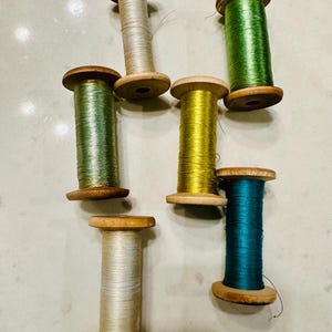 May include: Six wooden spools of thread arranged in a cluster. The threads are in various colors including light beige, sage green, bright lime green, golden yellow, teal, and a creamy off-white, all wound onto natural wood bobbins.