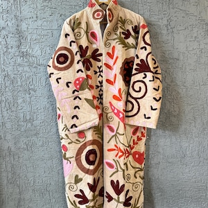 May include: A long, beige coat featuring intricate, colorful embroidery in shades of dark red, pink, and olive green. The design includes floral and circular patterns across the front and sleeves, with a high collar and a tag reading The Boho Hanger.