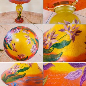 May include: A mushroom-shaped table lamp with a frosted yellow and orange glass base and shade. The lamp features hand-painted purple flowers with green leaves and a signature on the side. It includes a gold-toned metal hardware fitting.