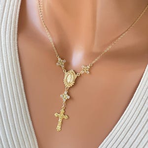 May include: A gold-toned rosary-style necklace featuring a delicate chain, small decorative floral accents, an oval-shaped pendant depicting a religious figure, and a textured cross charm hanging at the center of the piece.