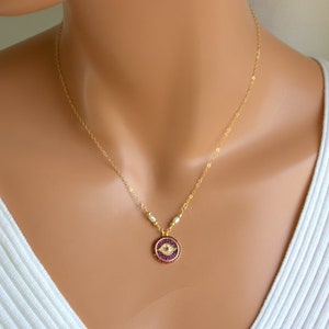May include: Gold chain necklace with a small round pendant featuring a red and purple evil eye design. The pendant is surrounded by small white pearls.