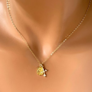 May include: A delicate gold-colored necklace with a small pendant featuring a detailed design and a smaller charm with clear stones. The necklace is displayed on a person's skin, highlighting its elegant and subtle design.