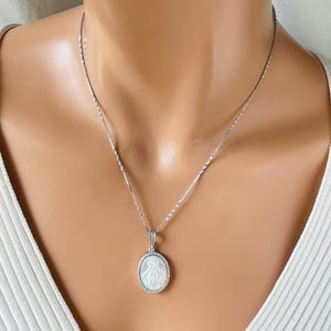 May include: A silver necklace featuring a pendant with a religious figure. The oval pendant has a white background and is surrounded by small, clear stones. The necklace is worn on a person wearing a white top.
