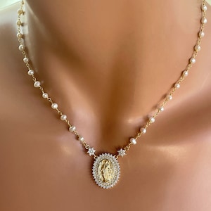 May include: Gold-toned necklace featuring a pendant with a religious figure surrounded by a halo of small, clear stones. The pendant is connected to a chain of small pearls. The necklace is displayed on a mannequin.