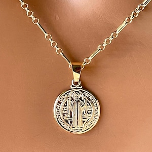 May include: A gold-colored necklace with a round pendant. The pendant features an embossed design with religious imagery and text. The chain is a delicate link style, and the necklace is displayed against a neutral background.
